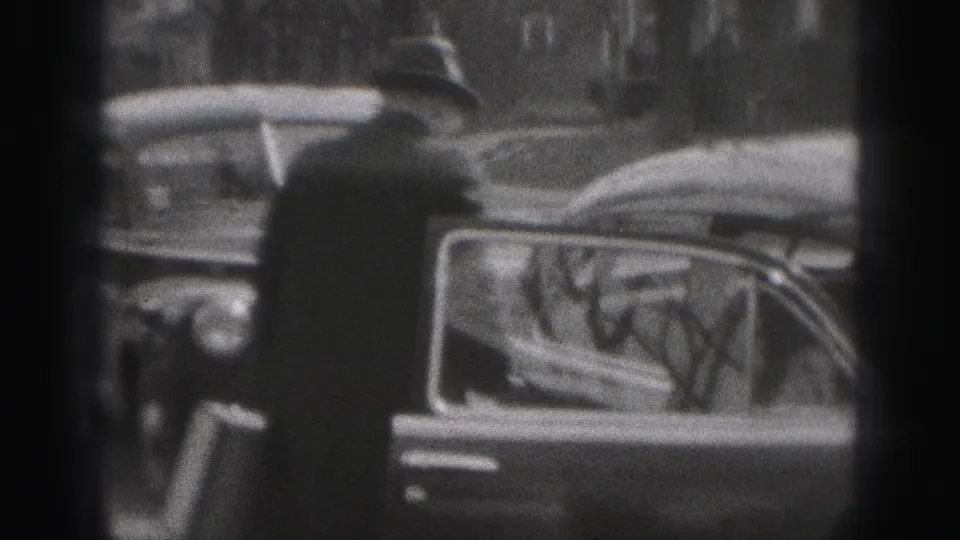 A man in a fedora and dark coat stands near a parked vintage car on the New Cumberland street in black-and-white 8mm footage.