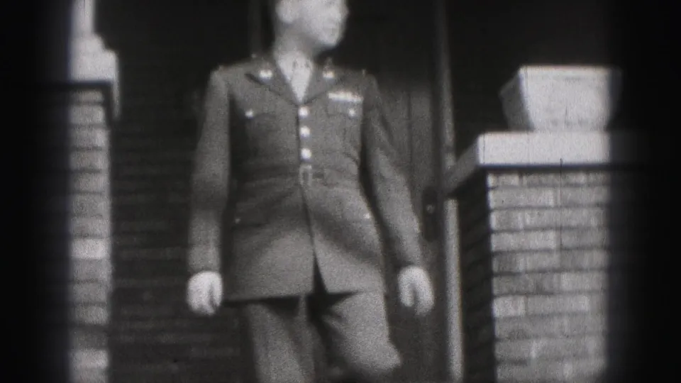 The man in formal military uniform continues down the house steps, partly soft from the 8mm home-movie focus.