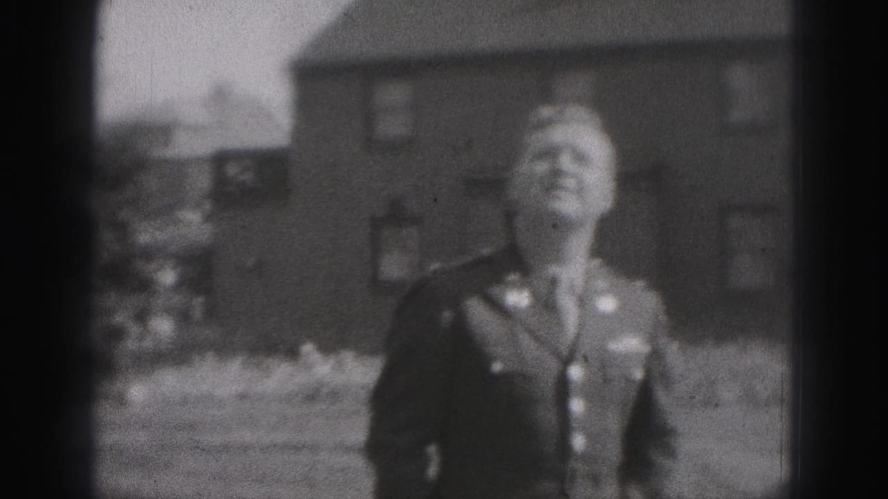 A man in military uniform stands outdoors near brick buildings in New Cumberland, smiling as the handheld 8mm camera frames him from a distance.