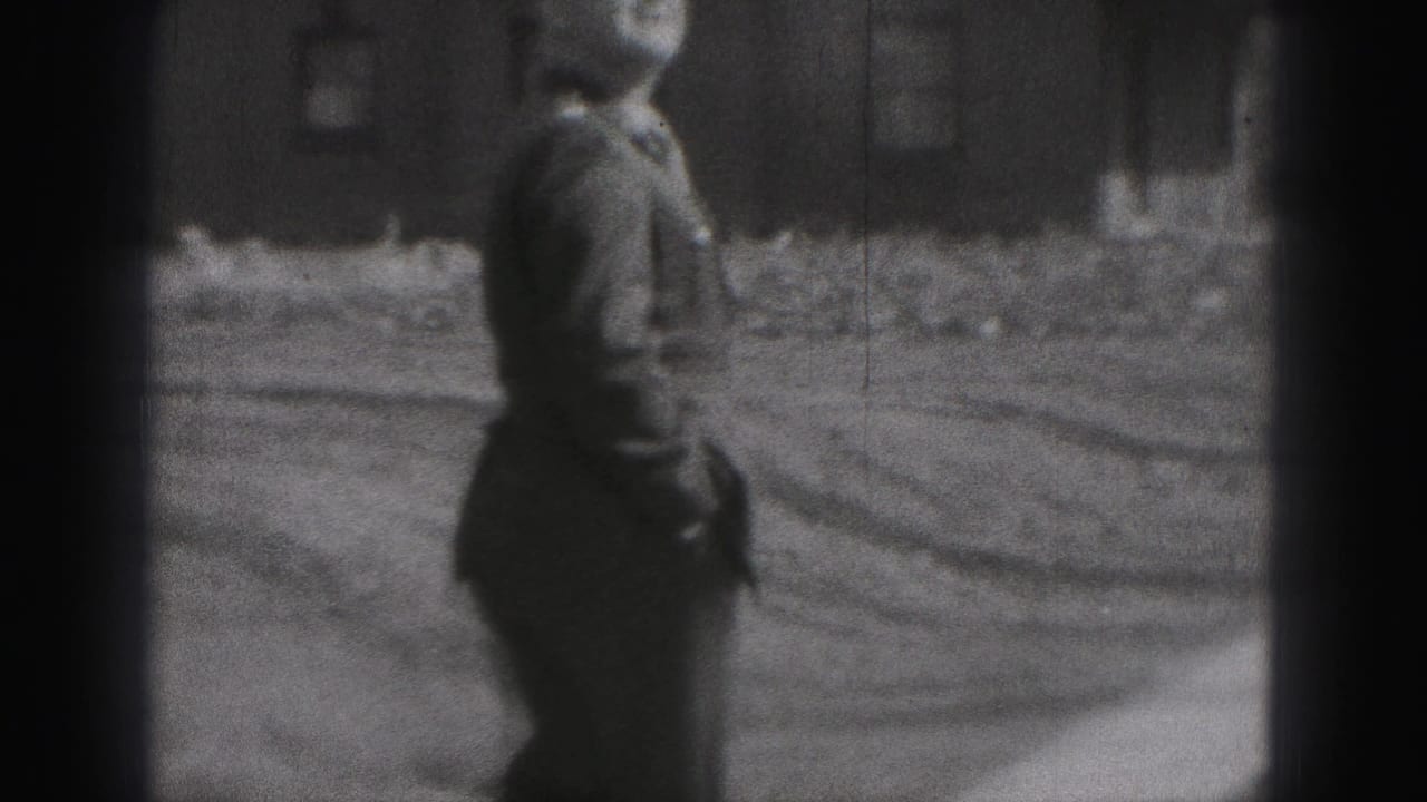 The uniformed man turns in profile and walks across the outdoor yard, with brick buildings and a path or road behind him.