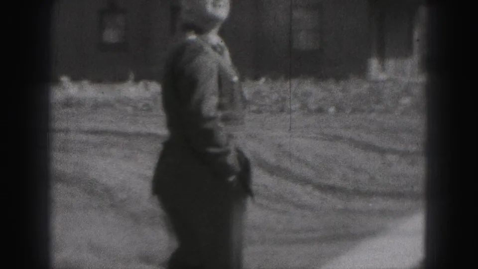 The uniformed man turns in profile and walks across the outdoor yard, with brick buildings and a path or road behind him.