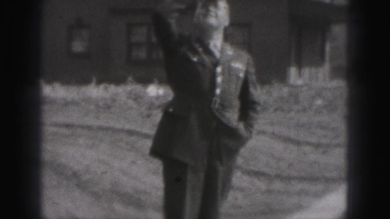 The uniformed man walks through the yard and smiles in a black-and-white 8mm frame, with simple brick buildings in the background.