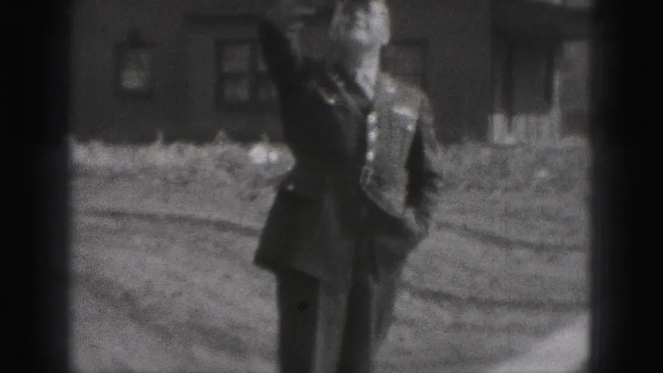 The uniformed man walks through the yard and smiles in a black-and-white 8mm frame, with simple brick buildings in the background.