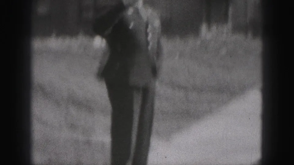 In 1946 New Cumberland home-movie footage, a man in dark formal clothing walks beside a grass verge and residential building.