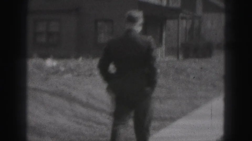The man is filmed from behind as he continues along a New Cumberland sidewalk past a residential facade in soft black-and-white 8mm footage.