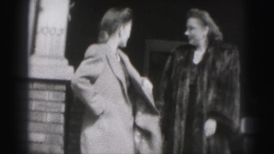 In 1946 New Cumberland footage, two women in coats stand close together outside a brick building, smiling as they turn toward each other.