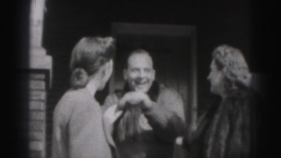 The 1946 home-movie frame shows a smiling man between two women by a doorway, with the group turned toward one another in conversation.