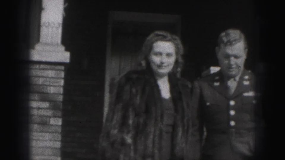 A woman in a fur coat stands beside a man in military uniform outside a brick building in 1946 New Cumberland footage.