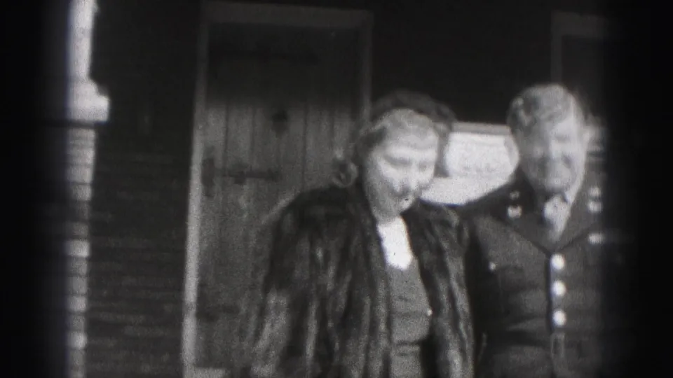 In 1946 New Cumberland, Pennsylvania footage, a uniformed man and a woman in a fur coat stand close together by a brick house entrance.