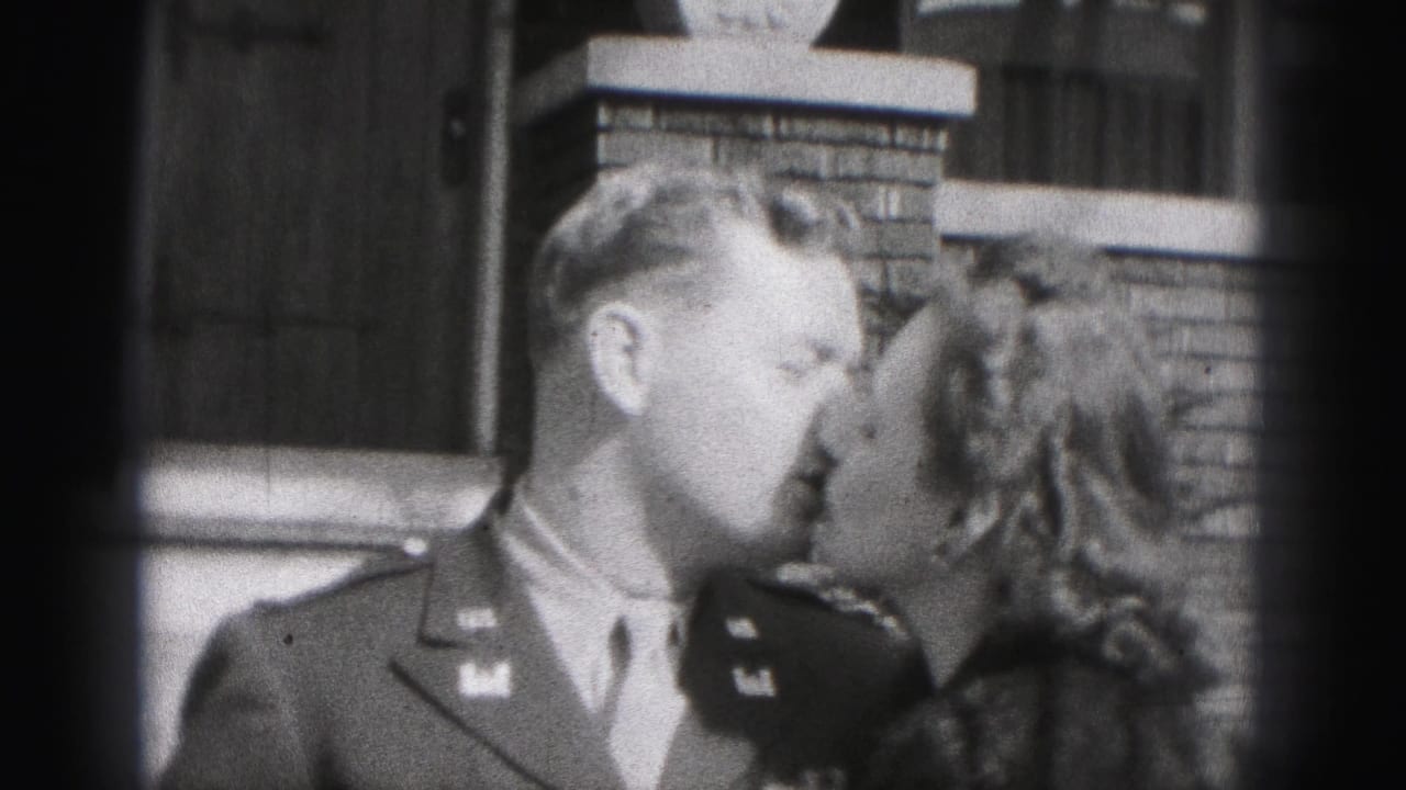 In the 1946 New Cumberland home-movie frame, a man in military uniform kisses a woman in a dark coat with a fur collar beside a brick house exterior.