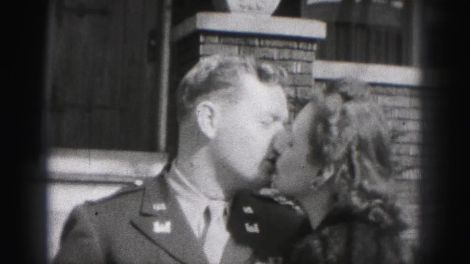In the 1946 New Cumberland home-movie frame, a man in military uniform kisses a woman in a dark coat with a fur collar beside a brick house exterior.