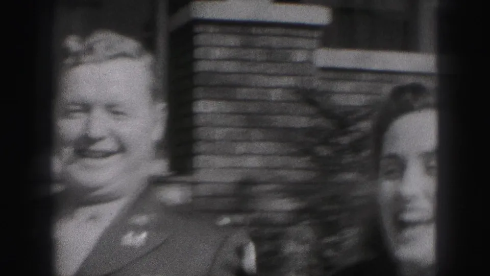 A smiling uniformed man and woman stand close together in front of a brick house wall in this 1946 New Cumberland home-movie frame.