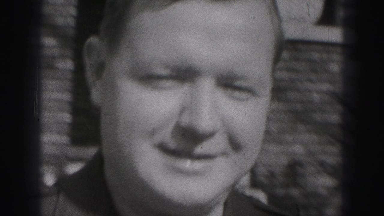 Close black-and-white frame of the smiling uniformed man in front of the brick house background, with the woman partly visible at the side.