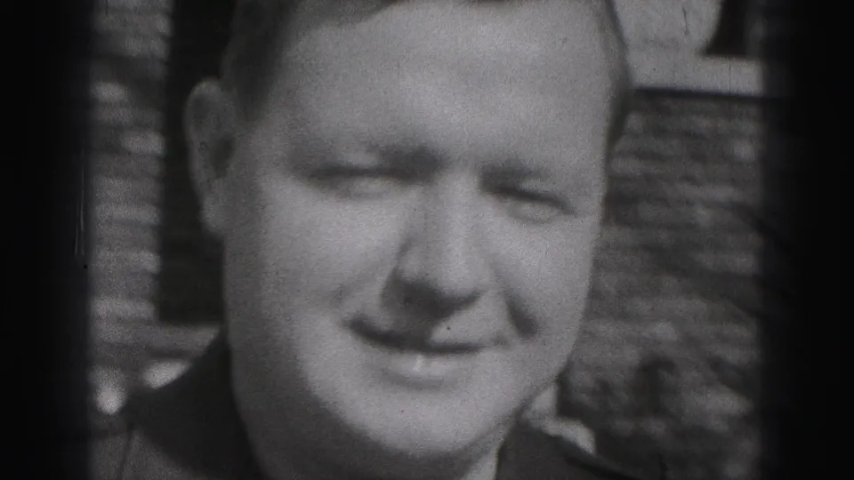 Close black-and-white frame of the smiling uniformed man in front of the brick house background, with the woman partly visible at the side.