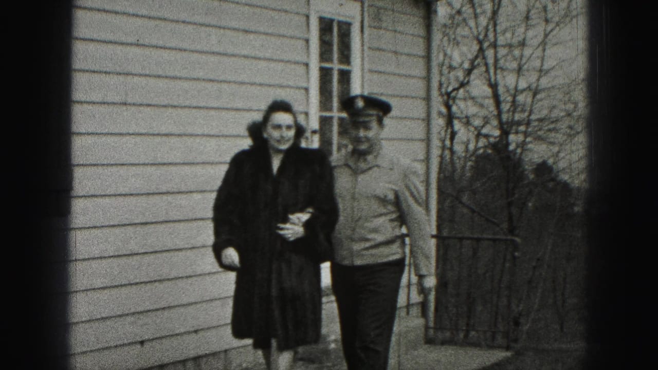Woman in dark coat walks arm-in-arm with a uniformed man beside a clapboard house.