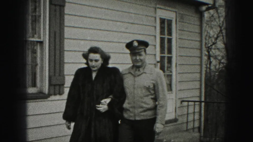 The woman and uniformed man continue walking arm-in-arm outside the New Cumberland house, with bare trees and siding behind them.