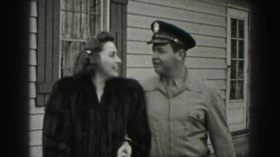 The woman turns toward the uniformed man while they walk together outside the New Cumberland residence in black-and-white 8mm footage.