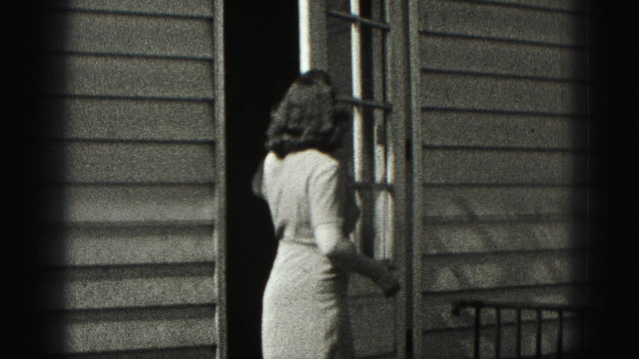 Woman in a light dress stands at a glass-paneled exterior door beside clapboard siding.