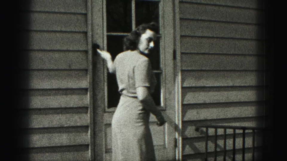 The woman stands outside the house door and looks toward the camera while holding or reaching near the door frame.