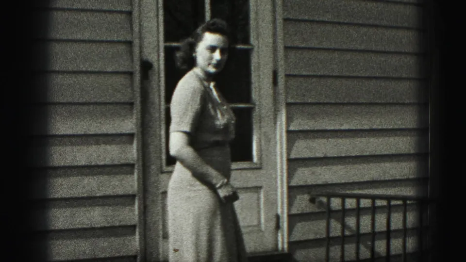 A woman in a mid-century dress stands beside the house door, with clapboard siding and porch railing visible behind her.