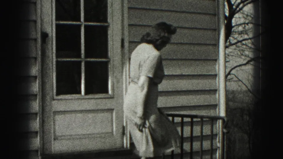 The woman moves away from the glass-paneled door along the small porch, with house siding and bare branches visible.
