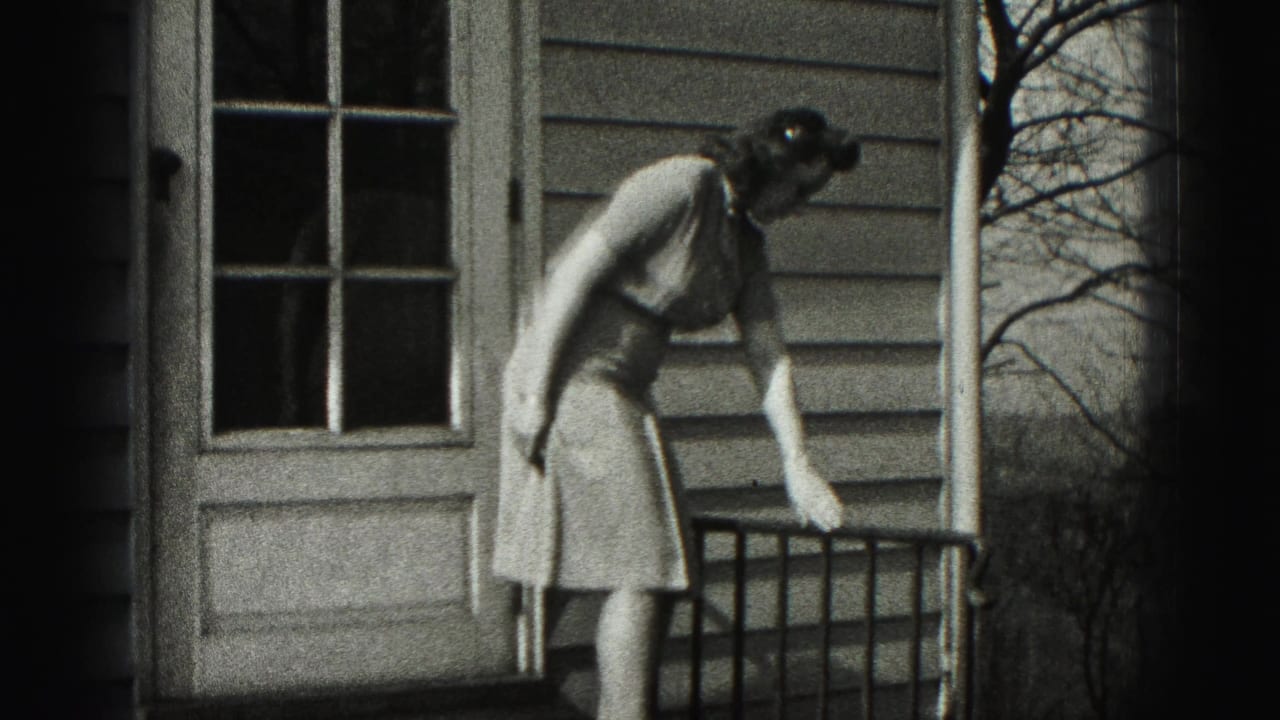 In 1946 New Cumberland home-movie footage, a woman in a light dress leans near a porch railing beside a clapboard house door.