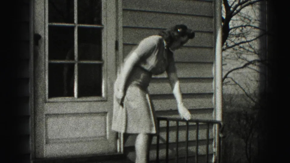 In 1946 New Cumberland home-movie footage, a woman in a light dress leans near a porch railing beside a clapboard house door.