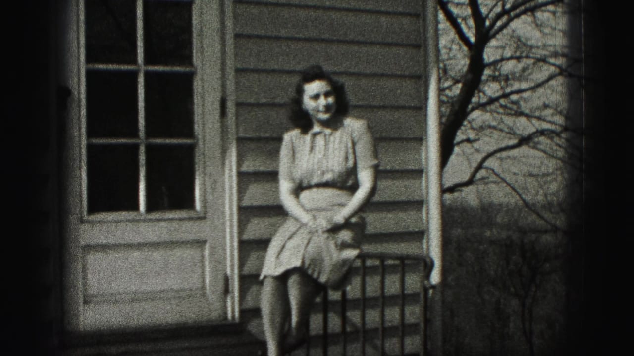 A woman in a 1940s-style dress sits facing the camera on a porch in New Cumberland, with house siding and a door behind her.
