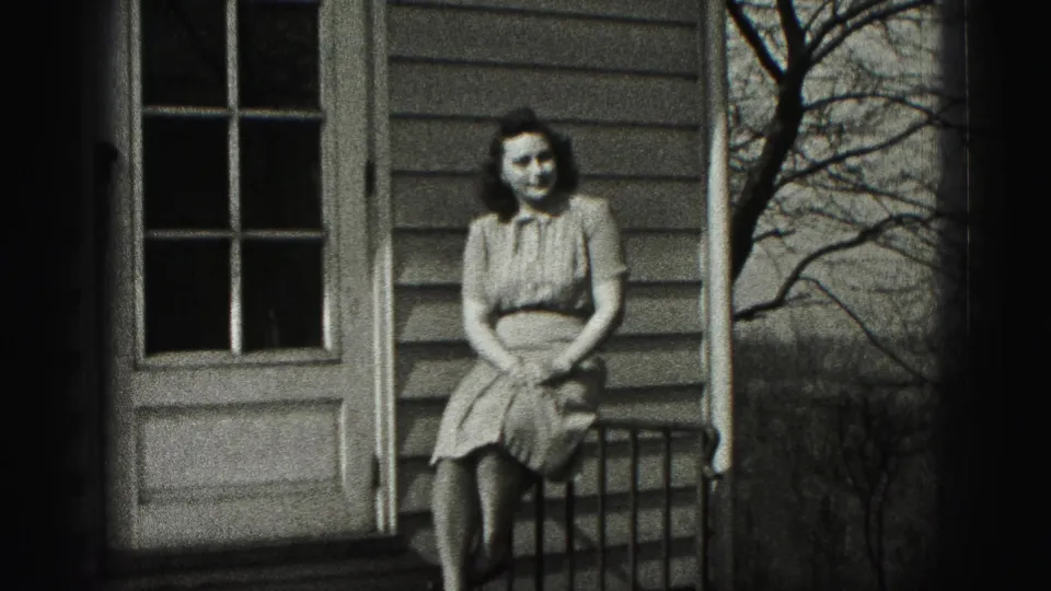 A woman in a 1940s-style dress sits facing the camera on a porch in New Cumberland, with house siding and a door behind her.