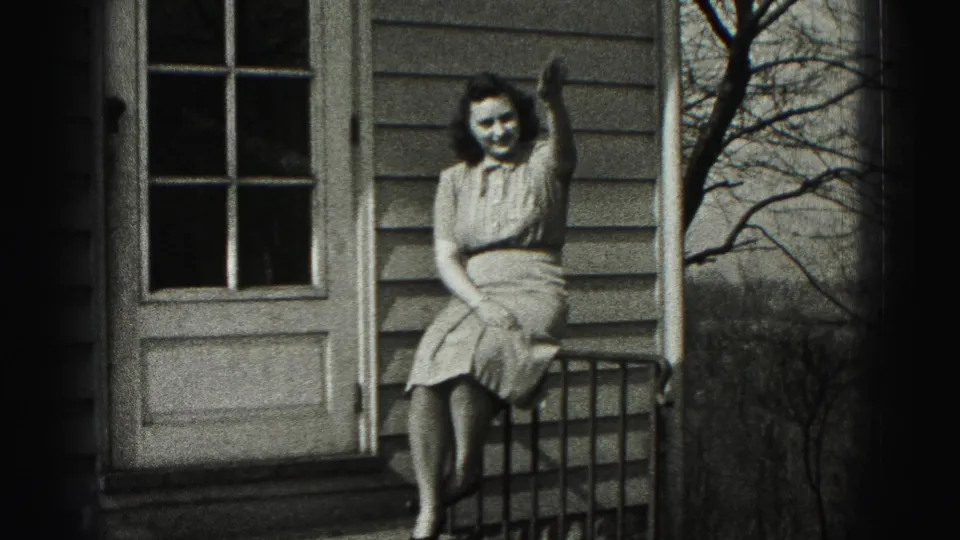 The seated woman smiles and waves toward the camera from a residential porch in 1946 New Cumberland home-movie footage.