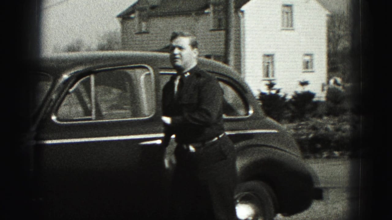 Man in a dark suit stands by a rounded 1940s sedan in a residential New Cumberland setting.