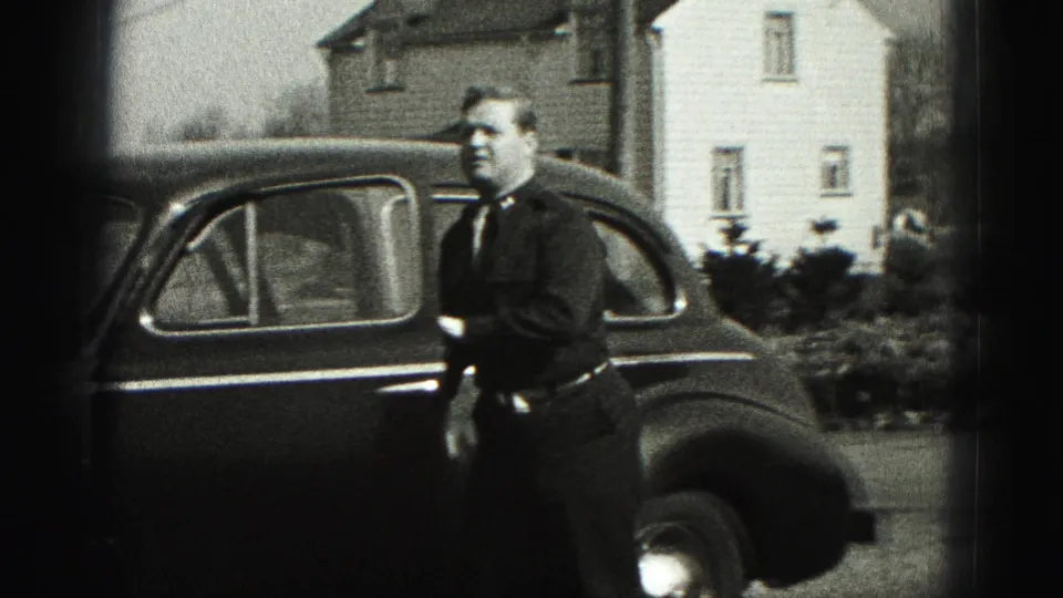 Man in a dark suit stands by a rounded 1940s sedan in a residential New Cumberland setting.