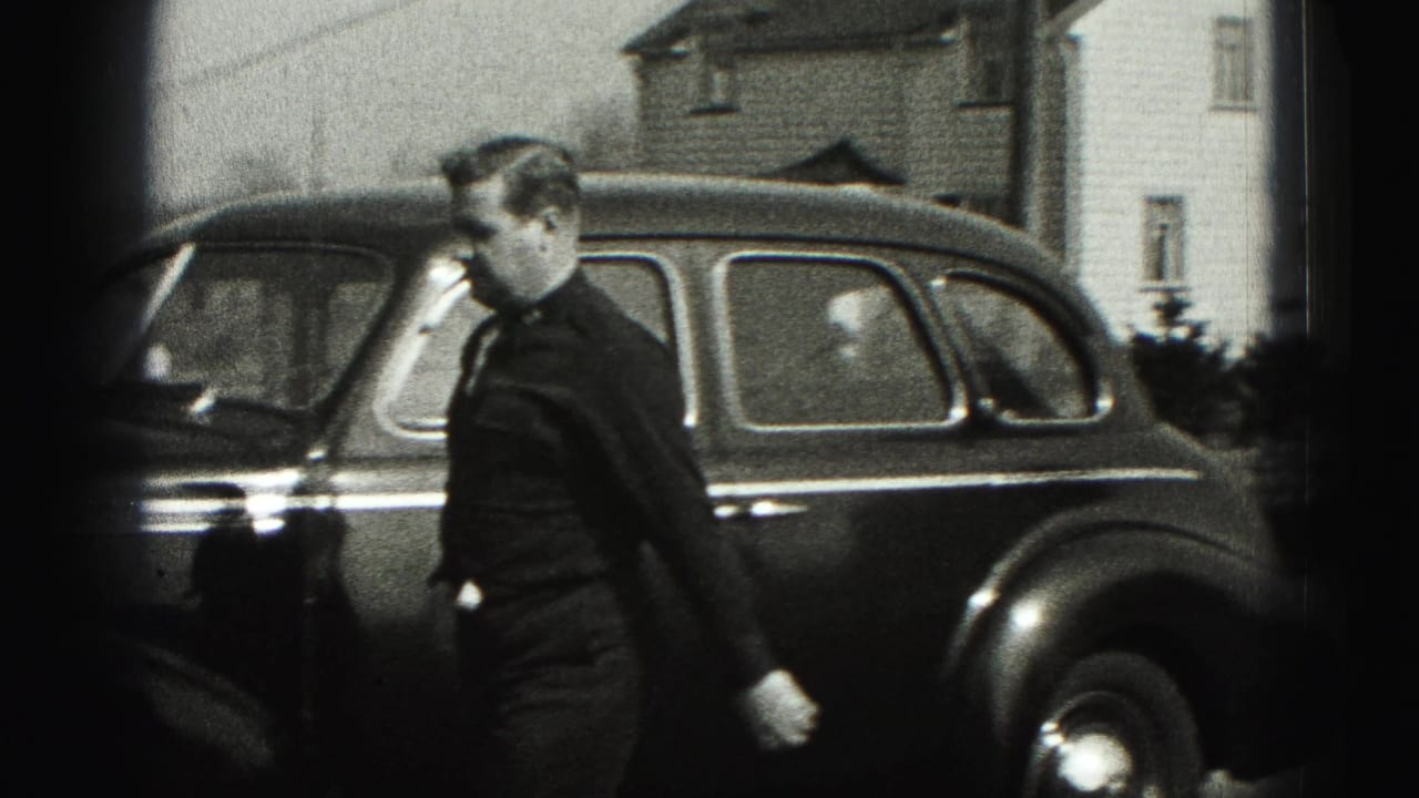 Man in a dark suit walks past a rounded sedan outside houses in the 1946 New Cumberland home movie.