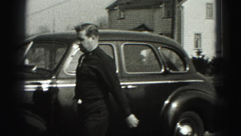 Man in a dark suit walks past a rounded sedan outside houses in the 1946 New Cumberland home movie.