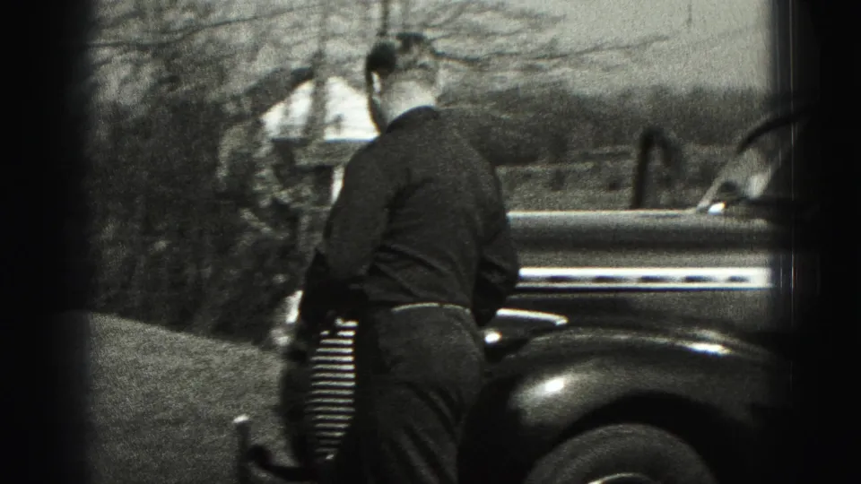 Man seen from behind approaches the front of a rounded sedan near a house and trees in New Cumberland.