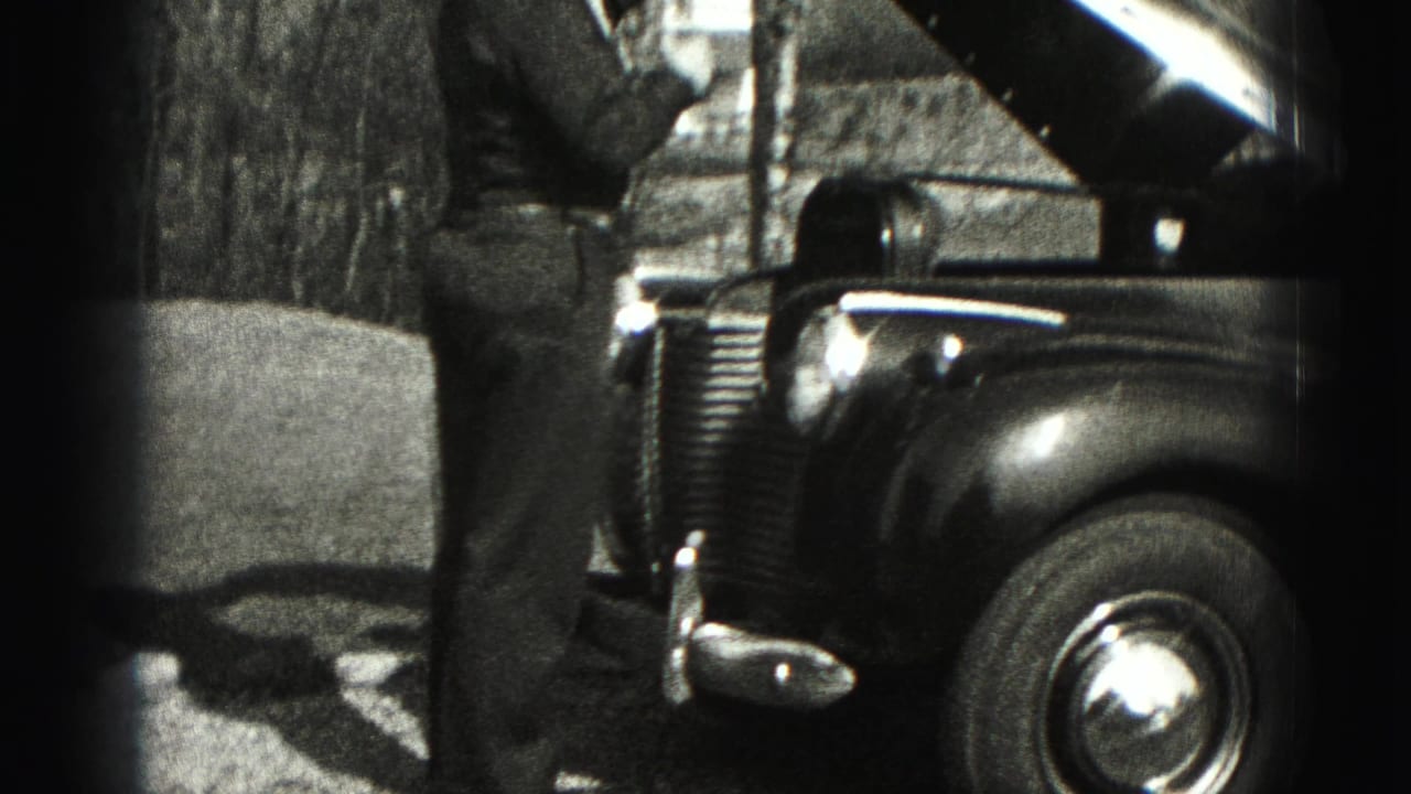 A man stands beside the open hood of a 1940s sedan, with the front wheel, grille, and trees visible.