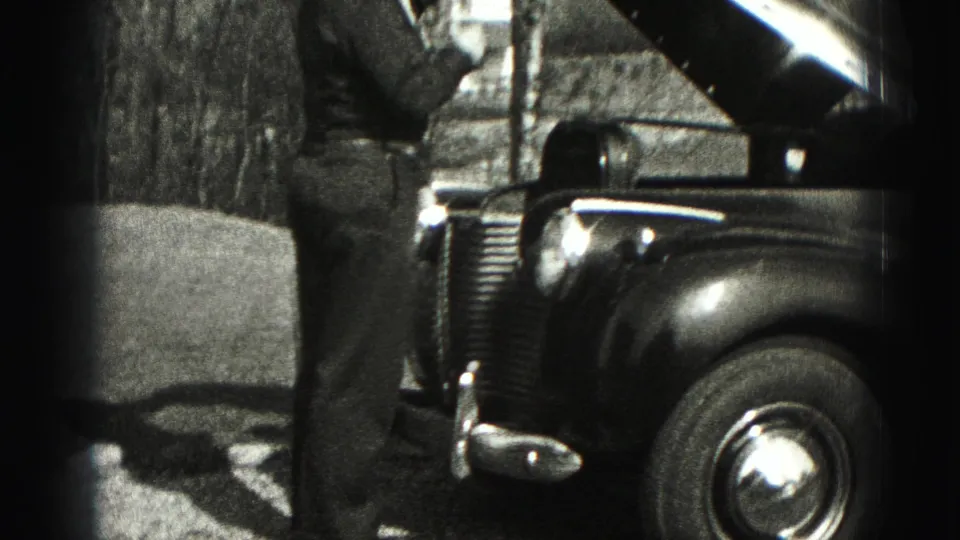 A man stands beside the open hood of a 1940s sedan, with the front wheel, grille, and trees visible.