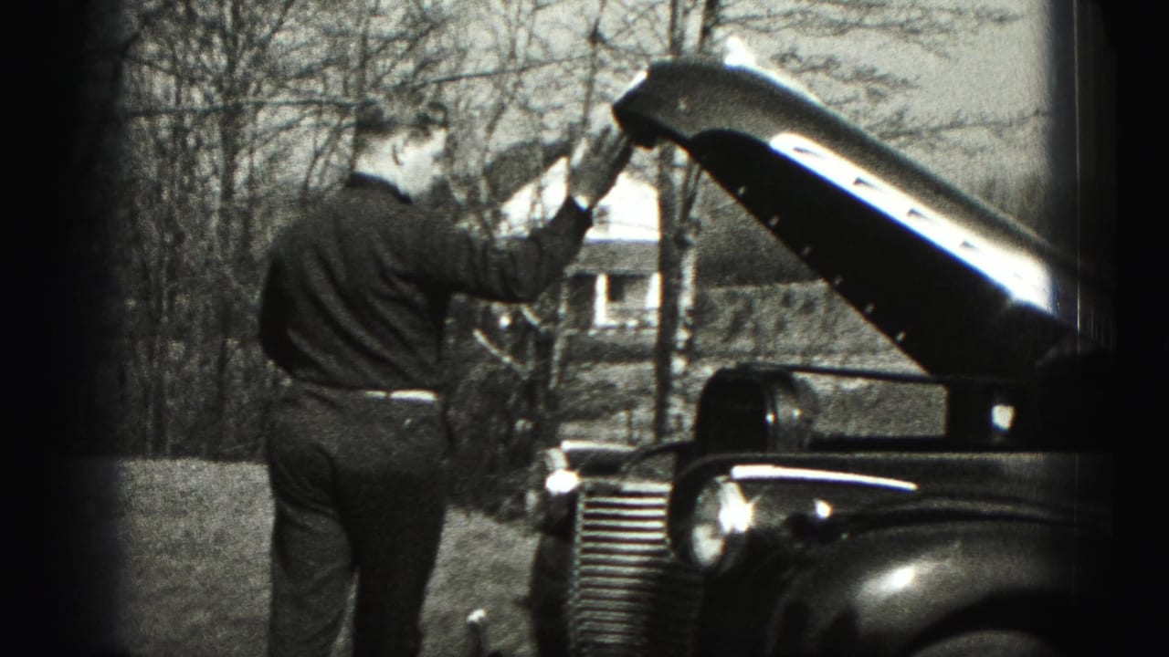 A man in formal clothes holds the open hood of a vintage sedan in a rural New Cumberland setting.