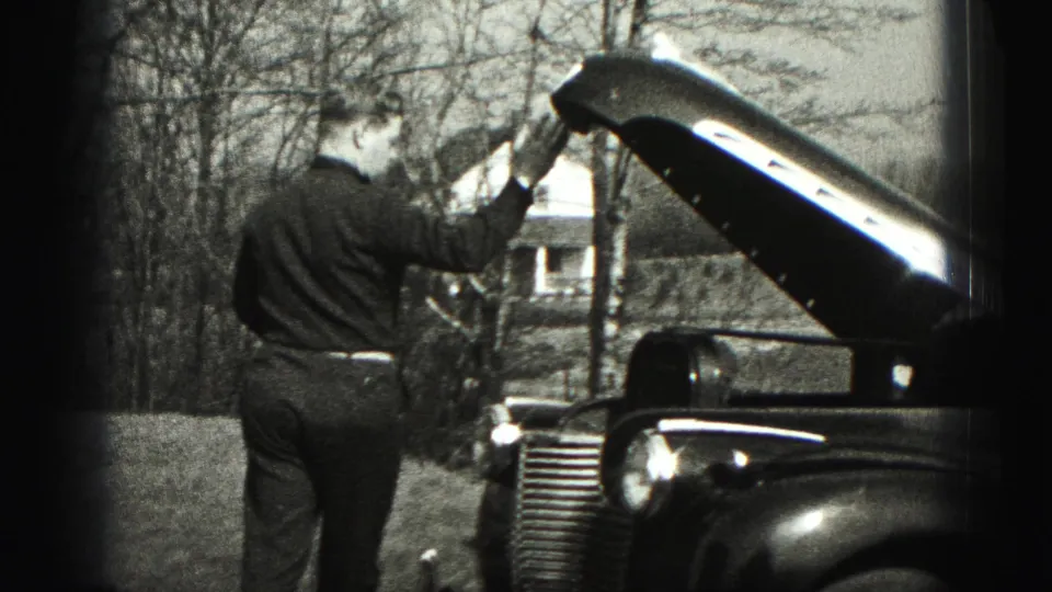 A man in formal clothes holds the open hood of a vintage sedan in a rural New Cumberland setting.