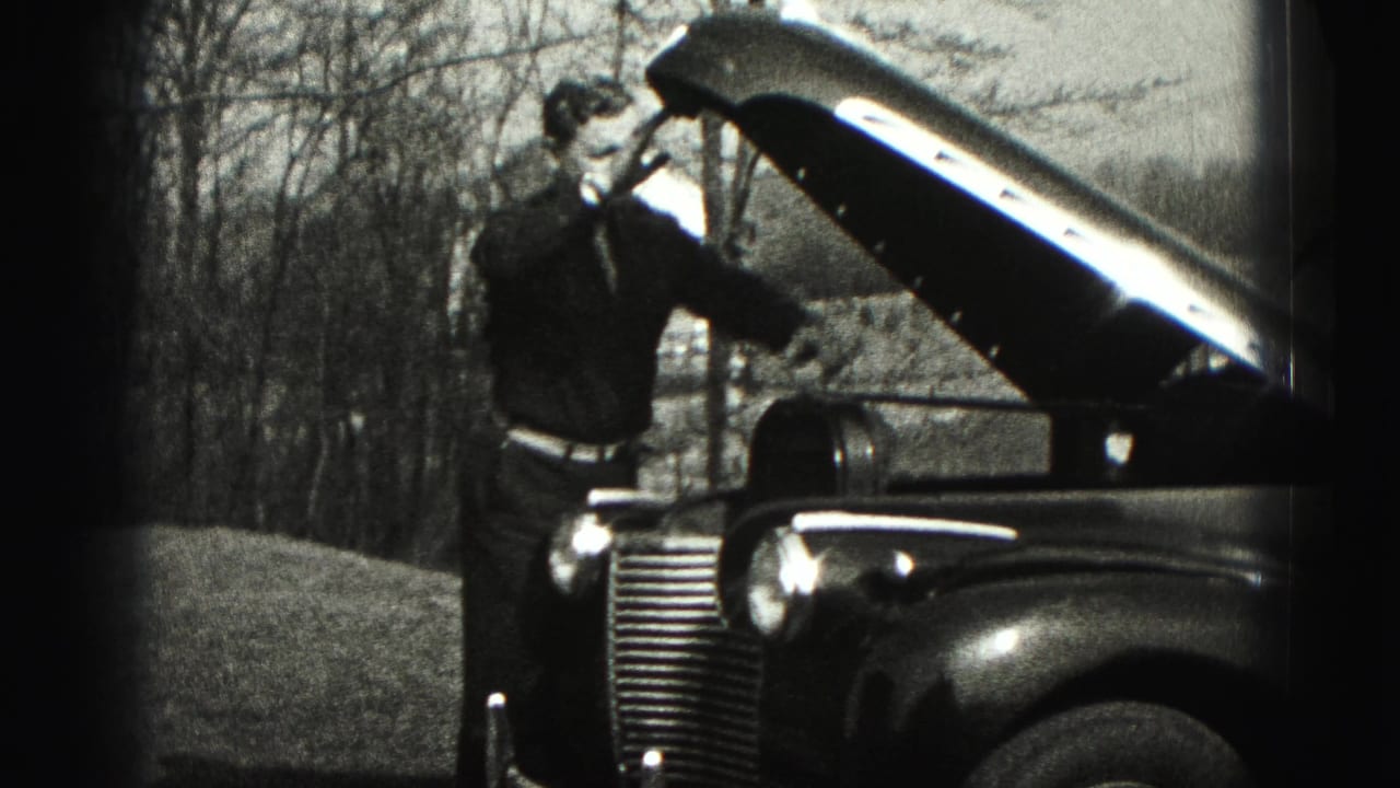 The man stands at the open hood of the 1940s sedan, framed by bare trees and rural ground.