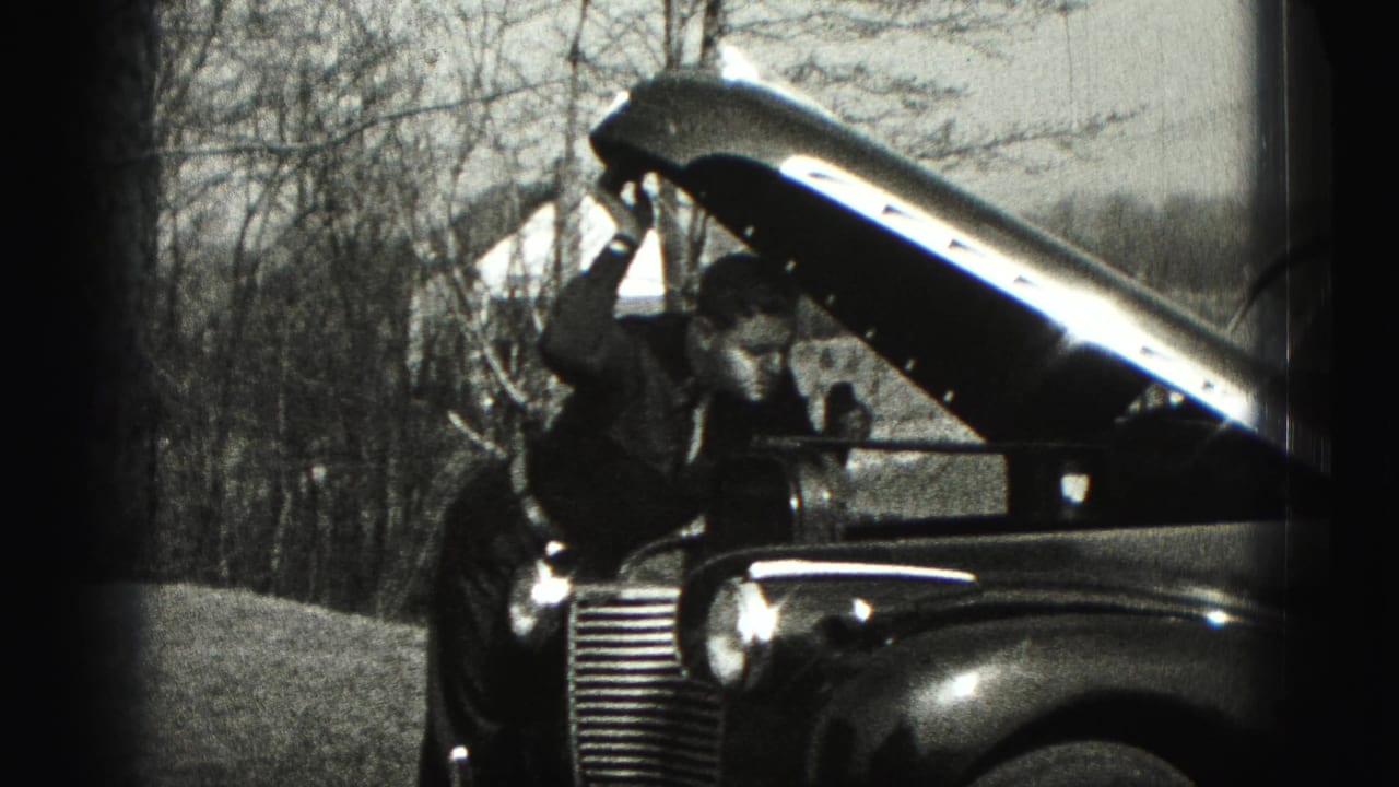 A man leans forward into the open hood area of a vintage automobile during the 1946 outdoor home-movie scene.
