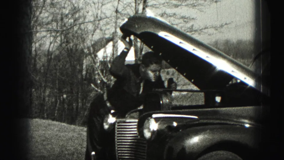 A man leans forward into the open hood area of a vintage automobile during the 1946 outdoor home-movie scene.