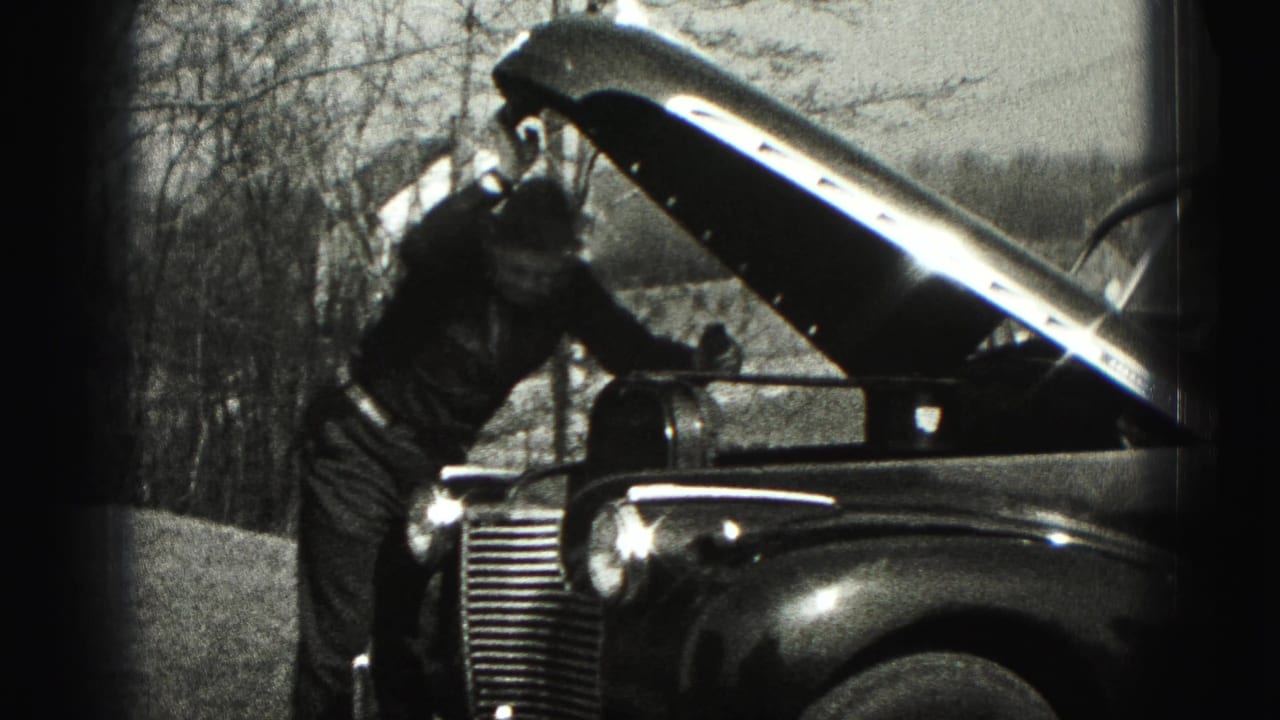 The man remains beside the open hood of the parked vintage car, partly turned toward the engine area outdoors.