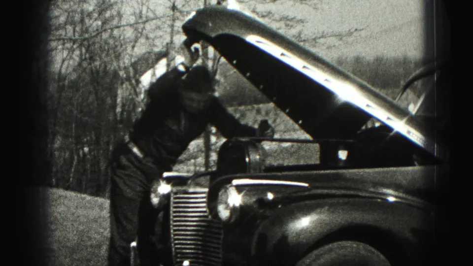 The man remains beside the open hood of the parked vintage car, partly turned toward the engine area outdoors.