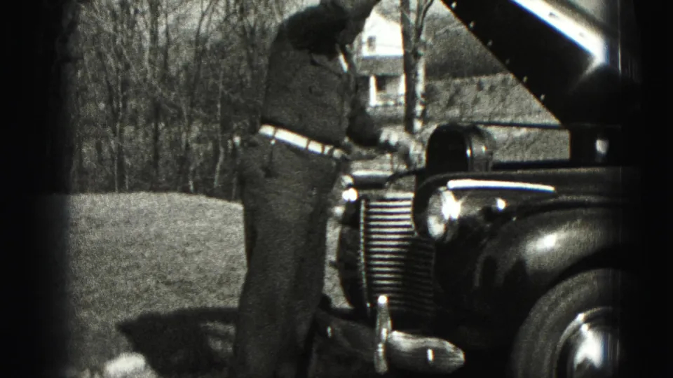 A suited man reaches down into the engine area of a vintage car, with an open hood and grassy yard visible behind him.