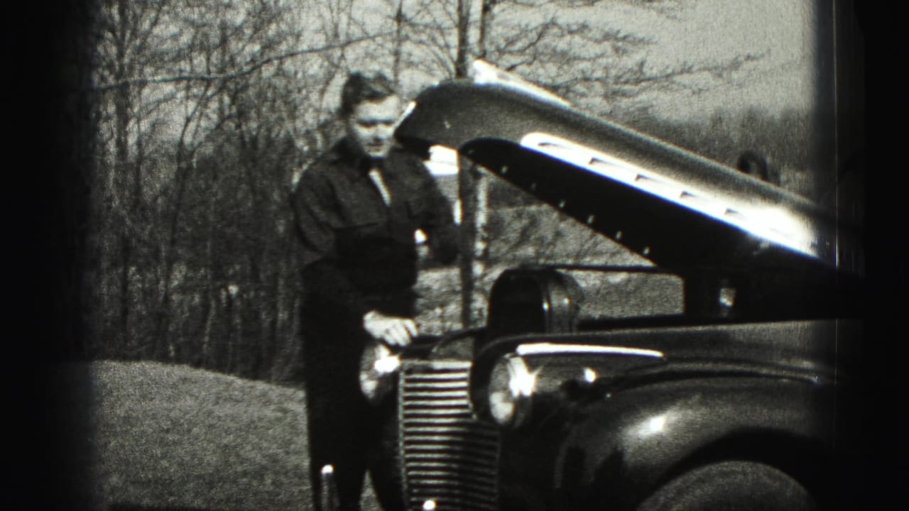 Man stands beside a classic car with its hood open in a yard.