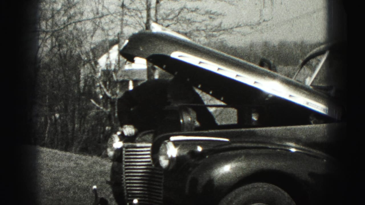 A man bends over the open engine bay of a 1940s car in a New Cumberland yard, with bare trees behind him.