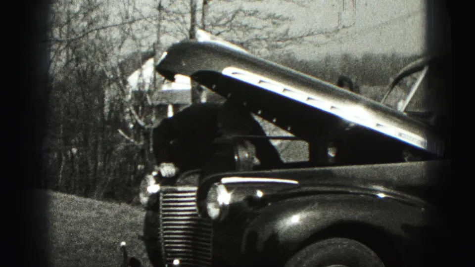 A man bends over the open engine bay of a 1940s car in a New Cumberland yard, with bare trees behind him.