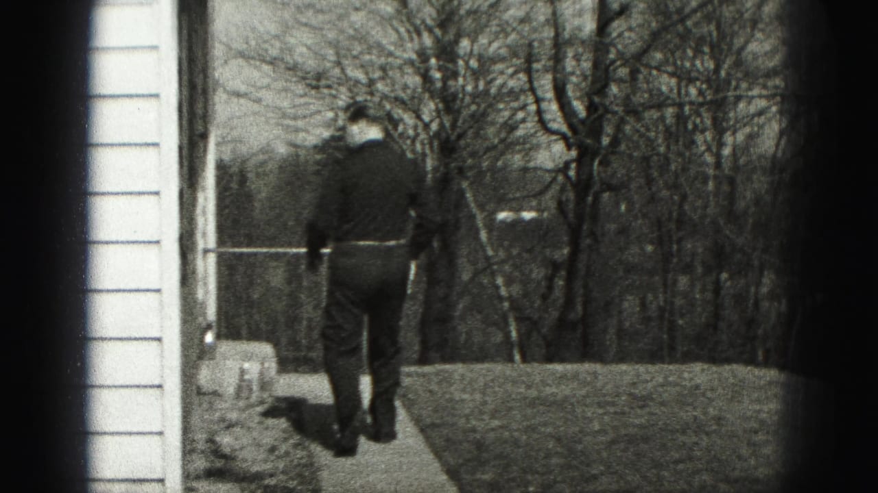 The man walks away along a narrow walkway beside house siding, with bare trees and lawn visible beyond.