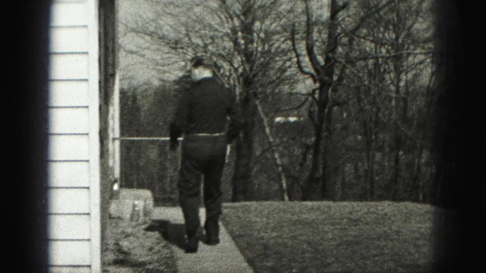 The man walks away along a narrow walkway beside house siding, with bare trees and lawn visible beyond.